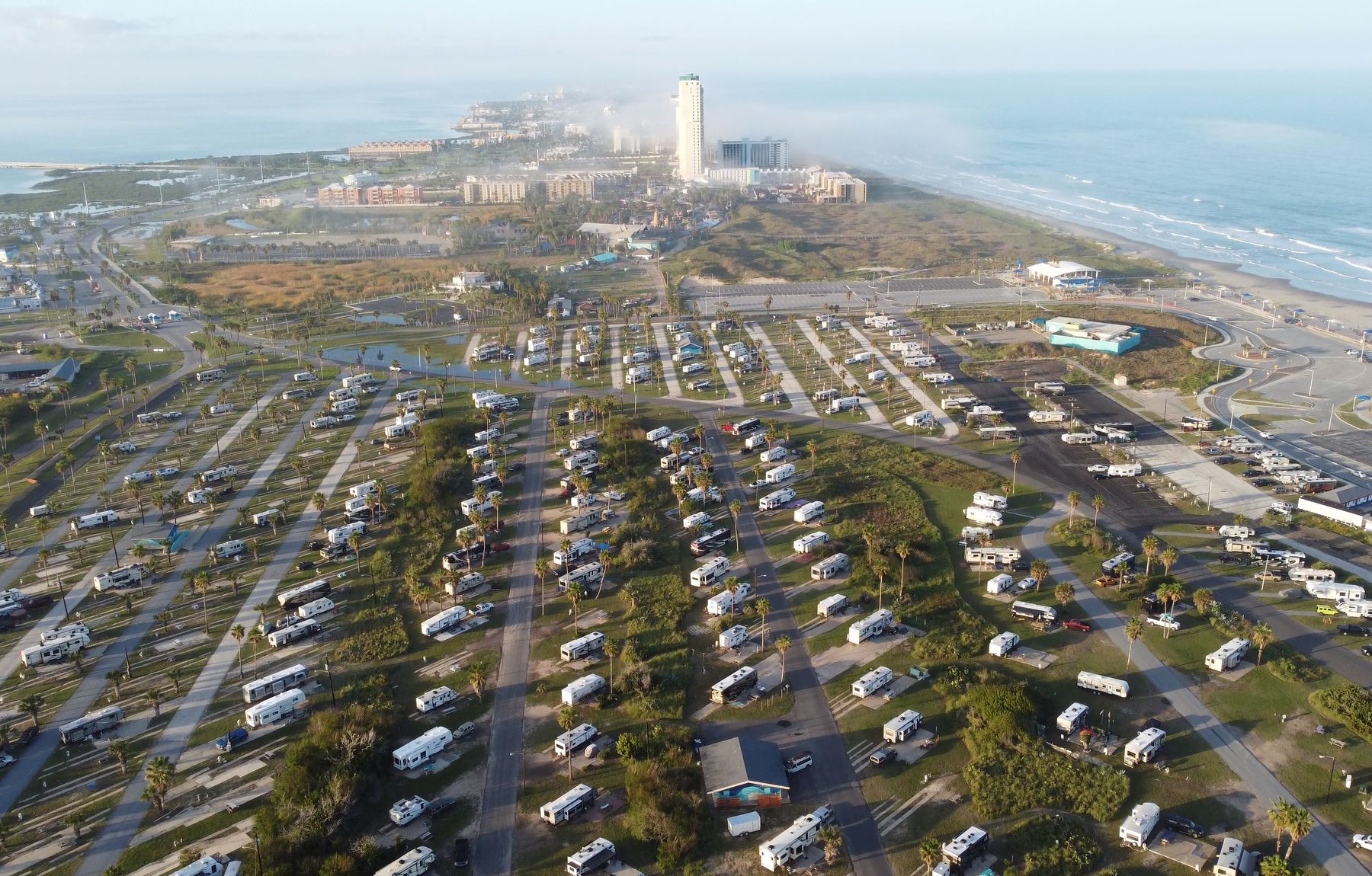 Aerial view of a large coastal RV resort, with hundreds of RV sites laid out in rows among palm trees beside the beach