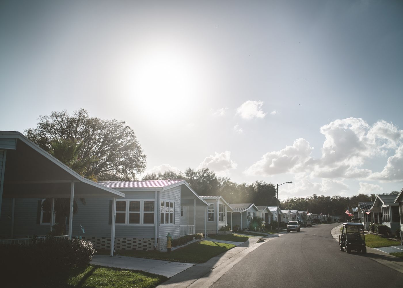 Street in a manufactured housing community lined with single-story homes, carports and American flags, a golf cart parked at the curb